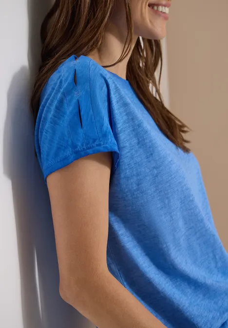 Nahaufnahme einer seitlichen lächelnden Frau in einem hellblauen Kurzarmshirt vor weißem Hintergrund.
