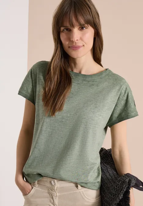 Jonge vrouw draagt groen T-shirt en beige broek, houdt losjes een jas over haar arm voor een lichte achtergrond.