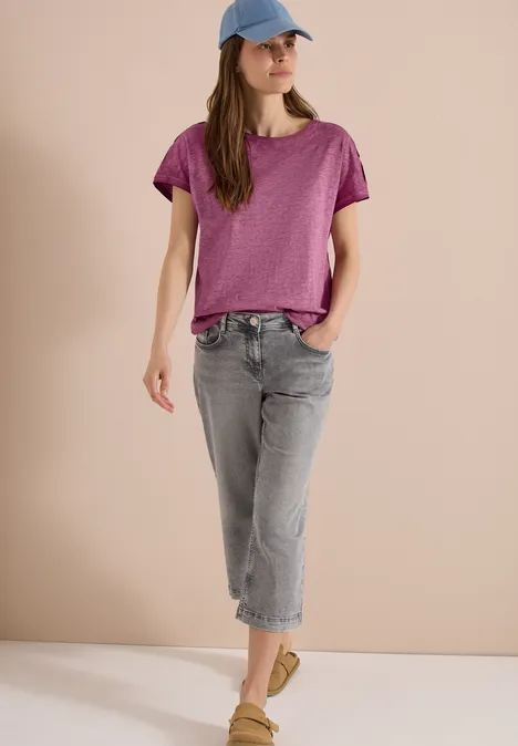 Person trägt lila T-Shirt, graue Jeans, grüne Schuhe und blaue Kappe vor beigem Hintergrund.