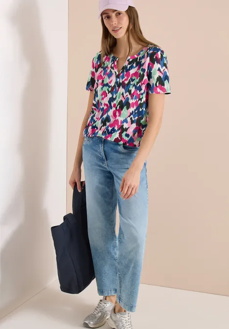 Een vrouw draagt een kleurrijke korte mouw blouse, een wijdvallende jeans, witte sneakers en houdt een donkerblauwe tas.