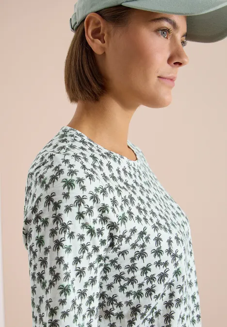 Opzij gezien draagt een vrouw een pet en een wit t-shirt met groen palmbomenpatroon.