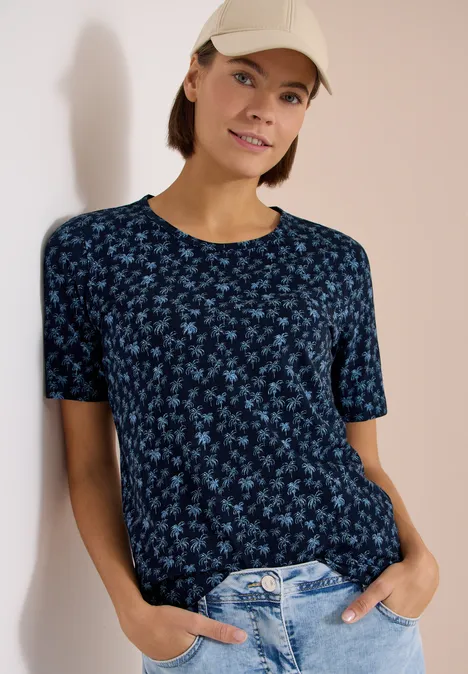 Vrouw leunt tegen muur, draagt donkerblauw bloemen-T-shirt, lichtblauwe jeans en pet, handen in zakken.