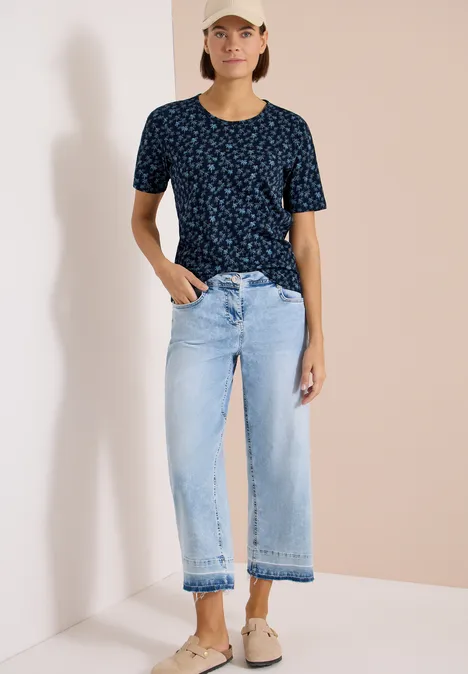Persoon in blauw T-shirt met print, lichtblauwe wijde cropped jeans en beige instappers.