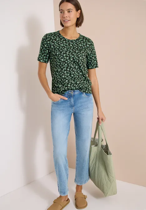 Vrouw met groen gebloemd T-shirt, lichtblauwe jeans, groene tas en bruine schoenen.