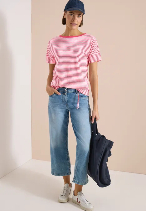 Eine stehende Person trägt rosafarben gestreiftes T-Shirt, weite Jeans und Sneakers, hält eine Jacke.