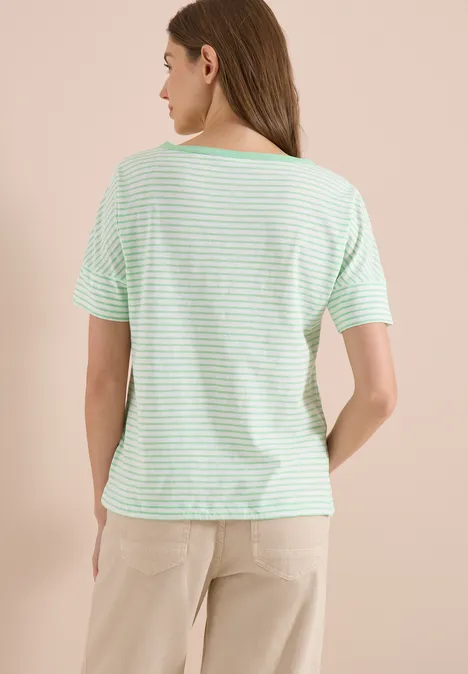 Eine Frau steht mit dem Rücken zur Kamera, trägt ein grün-weiß gestreiftes T-Shirt und helle Hose
