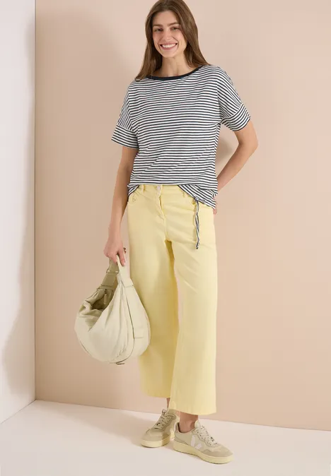 Vrouw in gestreept T-shirt, gele wijde culotte, lichtgroene tas en beige schoenen.