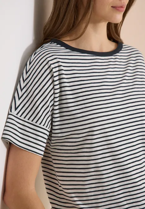 Vrouw leunt tegen een muur in een wit T-shirt met korte mouwen en marineblauwe strepen.