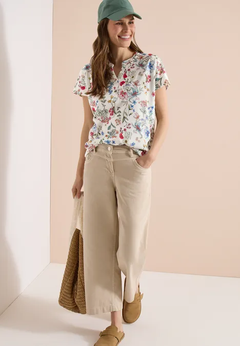 Frau in geblühmtem Shirt, hellen Culottes, Cap und Korbtasche vor neutralem Hintergrund.