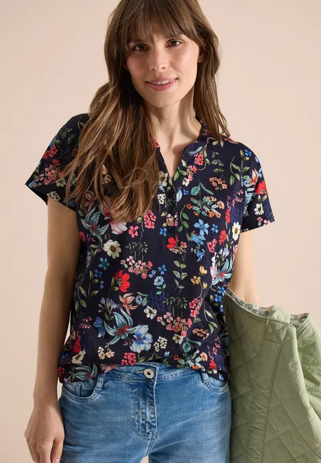 Vrouw draagt donkerblauw shirt met korte mouwen en bonte bloemenprint, een blauwe jeans en houdt een groene jas.