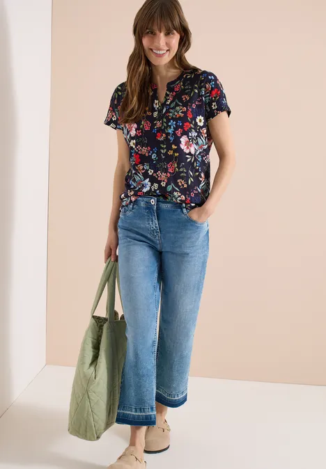 Vrouw in bloemen top, wijdvallende spijkerbroek, beige schoenen en groene tas tegen neutrale achtergrond.