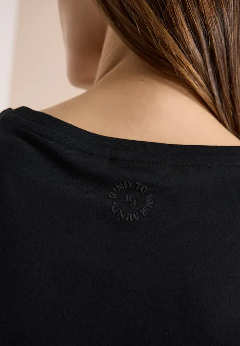 Rückansicht einer Frau mit langen Haaren in einem schwarzen Top, am Nacken ein kleines rundes Emblem.