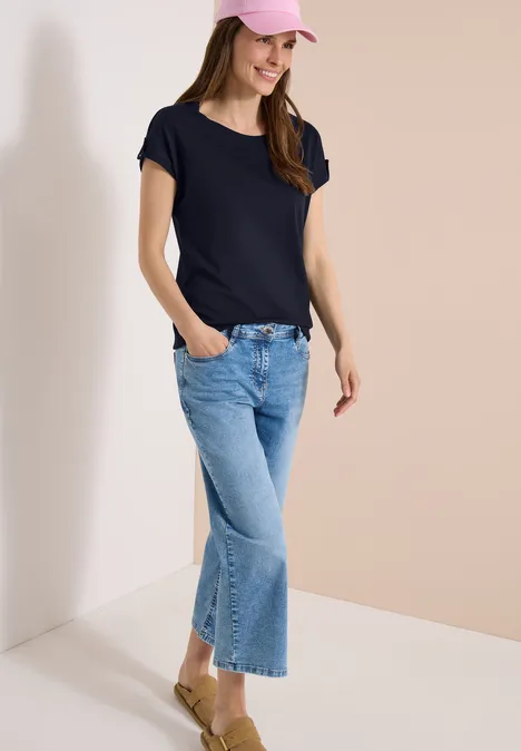 Frau in dunkelblauem T-Shirt und weitem, hellen Denim-Hosenrock, lässig mit Händen in den Taschen.