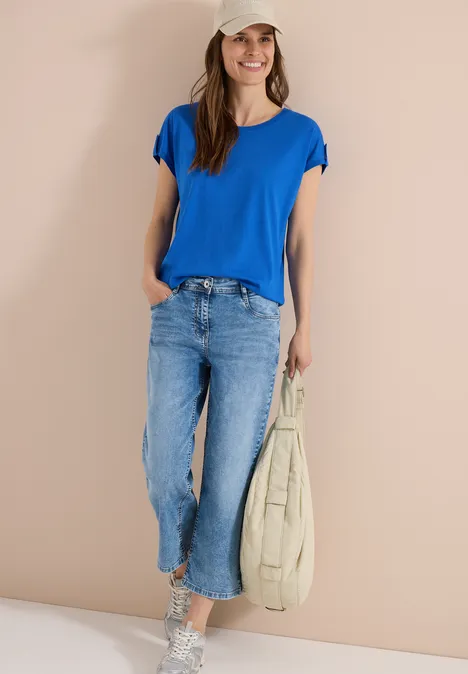 Frau in blauem T-Shirt und Jeans, hält eine beige Tasche, stehend vor neutralem Hintergrund.
