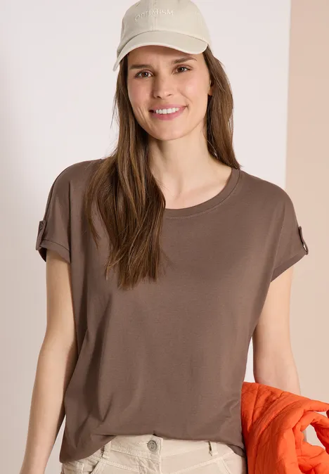 Frau in braunem T-Shirt und beiger Hose hält orange Jacke vor hellem Hintergrund.