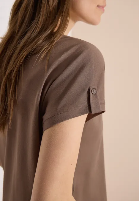 Seitliche Ansicht einer Frau in braunem Kurzarmshirt mit Knopf-Detail am Ärmel