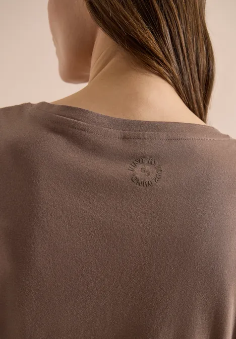 Nahaufnahme des Frauenrückens in braunem Shirt, Haar über eine Schulter, dezentes Logo am Nacken.