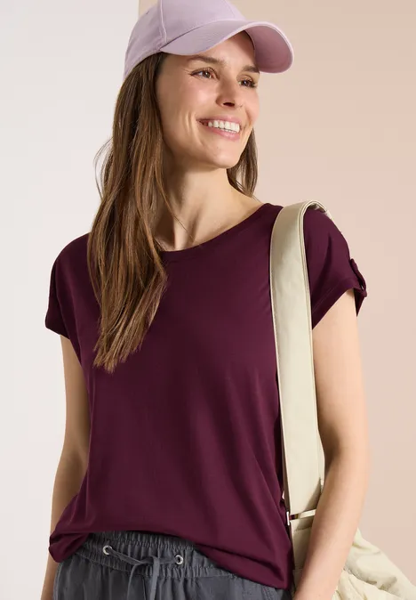 Frau mit burgunderfarbenem T-Shirt, lila Basecap und Schultertasche, Gesicht unscharf.