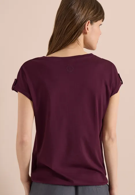 Frau von hinten in lockerem, violettem T-Shirt mit kurzen Ärmeln, vor beigem Hintergrund.