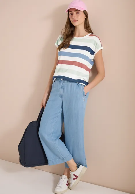 Frau in gestreiftem Shirt, hellblauen Culottes, weißen Sneakern und dunkelblauer Tasche vor beiger Wand.