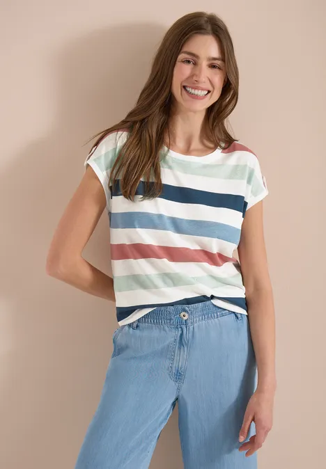 Eine Frau vor neutralem Hintergrund mit gestreiftem T-Shirt und hellen Jeans.