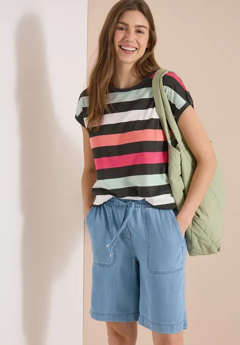 Person trägt gestreiftes T-Shirt, Jeans-Shorts und Rucksack vor neutralem Hintergrund.