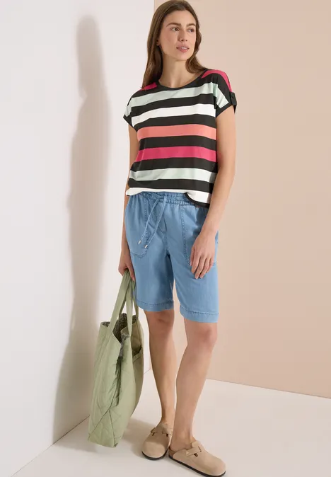 Eine Frau in bunt gestreiftem Shirt, hellblauen Bermudashorts und grüner Tasche.