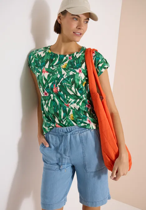 Frau in tropischem Print-Shirt, Jeansshorts, orangefarbener Stofftasche und Baseballcap.