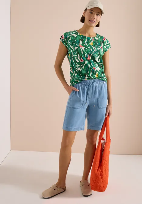 Frau mit grüner Blattmuster-Bluse, hellblaue Jeans-Shorts, beigen Clogs und orangefarbener Tasche vor beigem Hintergrund.
