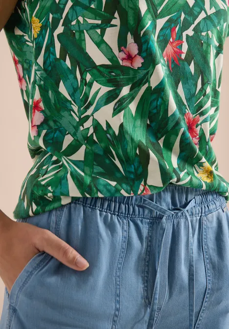 Person trägt grünes Top mit Blatt- und Blumenmuster und blaue Hose mit Gummizug, Hand in Tasche.