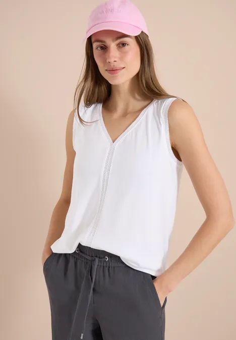 Vrouw met witte mouwloze top en grijze broek, handen in de zakken.