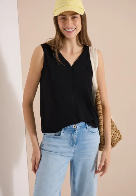 Femme en haut sans manches noir, jean clair et sac en paille tressé, posant devant un fond clair.