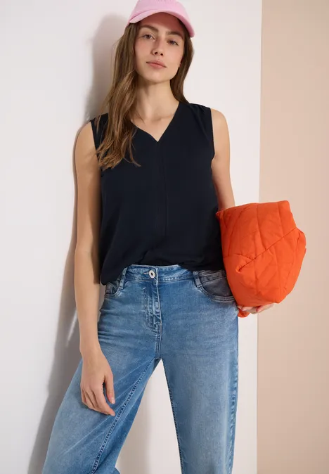 Vrouw in spijkerbroek en zwart topje houdt oranje kussen vast voor neutrale achtergrond.