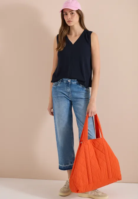 Vrouw met zwarte mouwloze top, blauwe cropped jeans, beige sneakers en rood doorgestikt tasje.
