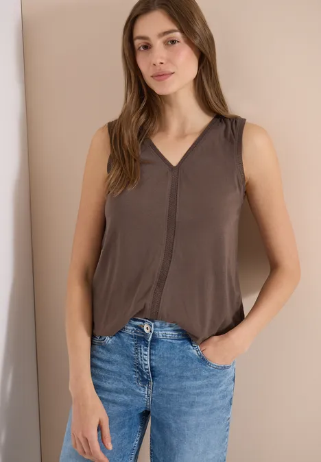 Frau in ärmellosem braunen Top und Jeans lehnt entspannt an der Wand mit einer Hand in der Tasche.
