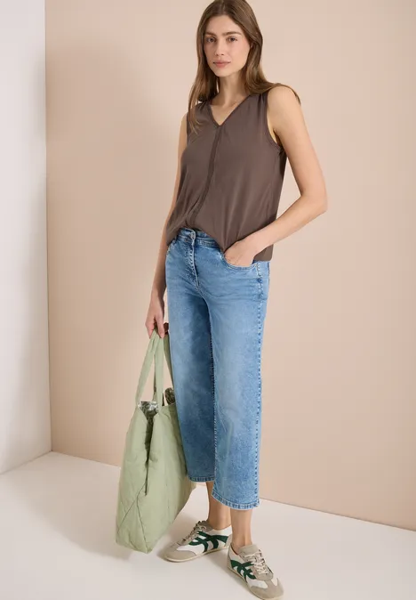 Frau mit braunem Top, weiten Jeans, grüner Tasche und Sneakern vor schlichtem, hellem Hintergrund.