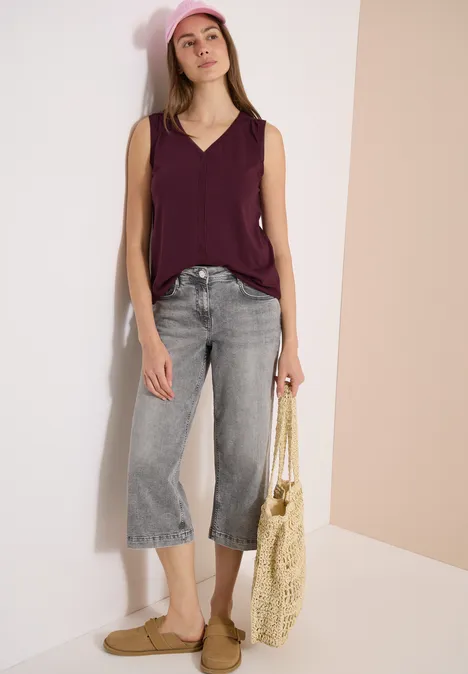 Vrouw in paars mouwloos topje, grijze wijde 7/8-jeans, strooien tas en platte sandalen voor een lichte muur.