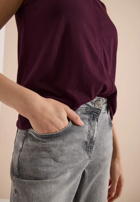 Persoon draagt een bordeauxrood hemd en grijze jeans, hand in voorzak.