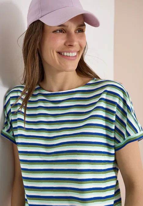 Ein Mädchen mit rosa Mütze und grün-blau gestreiftem T-Shirt lehnt an einer Wand.