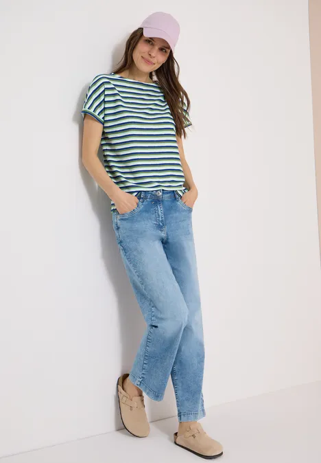 Eine Frau lehnt an einer Wand und trägt gestreiftes T-Shirt, Jeans und bequeme Schuhe.