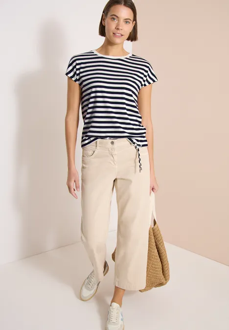 Vrouw in gestreept T-shirt, beige wijde broek, witte sneakers, met een strooien tas.