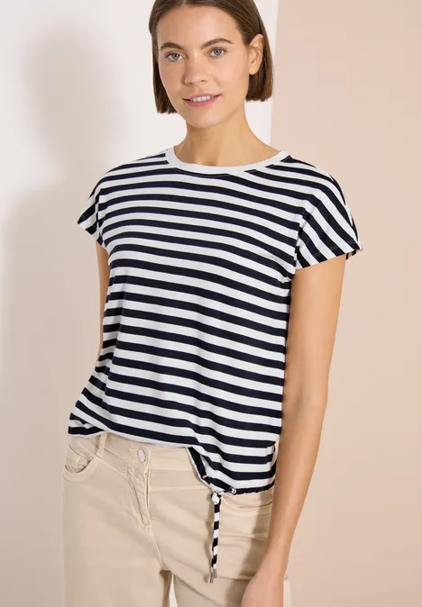 Vrouw in gestreept T-shirt en beige broek voor een neutrale achtergrond.
