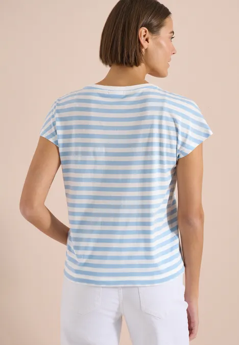 Rückenansicht einer Person in blau-weiß gestreiftem T-Shirt und weißer Hose vor beigem Hintergrund.