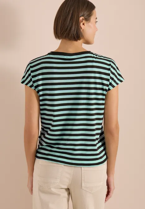 Person von hinten in schwarz-türkis gestreiftem T-Shirt und hellen Hosen vor beigem Hintergrund.