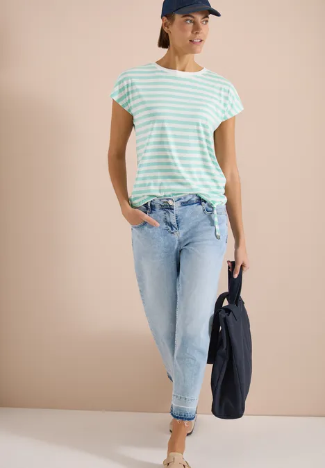 Dame in mintgrün-weiß gestreiftem Shirt, hellblauen Jeans mit Umschlag, hält eine dunkle Jacke.