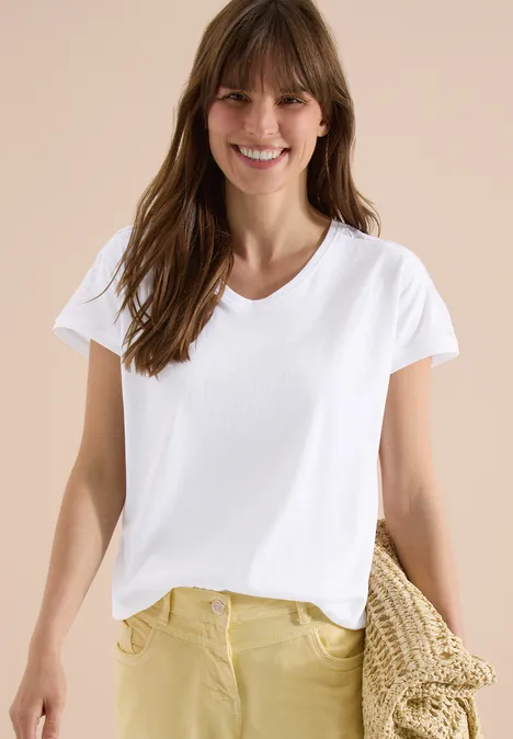 Frau mit weißen T-Shirt und gelber Hose hält einen geflochtenen Korb vor beigem Hintergrund.