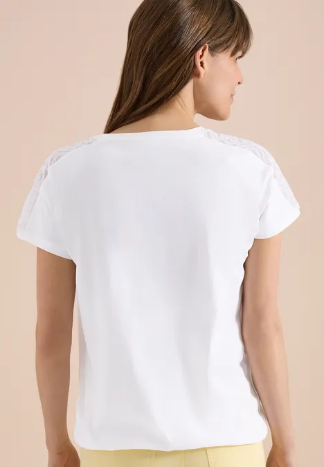 Eine Frau steht mit dem Rücken zur Kamera und trägt ein weißes T-Shirt mit Spitzenbesatz auf beigem Hintergrund.