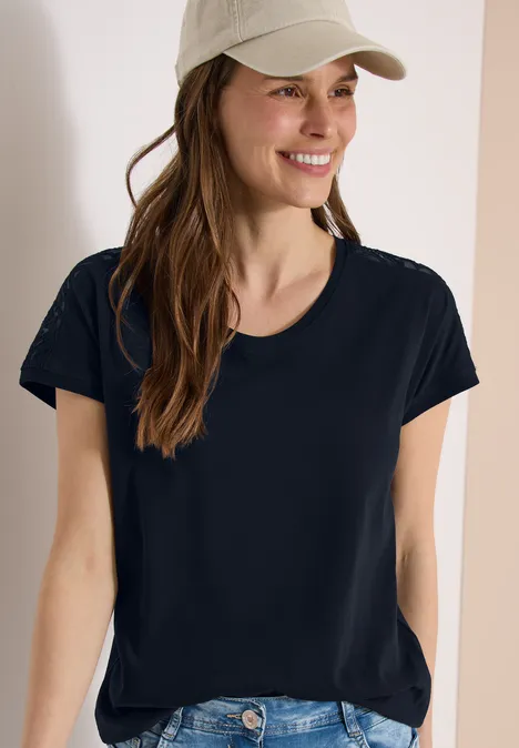 Frau mit schwarzem T-Shirt und Baseballcap, vor neutralem Hintergrund.