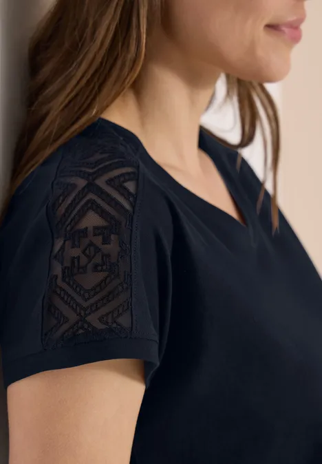 Eine Frau trägt ein schwarzes T-Shirt mit V-Ausschnitt und transparenten Spitzenärmeln mit geometrischem Muster.