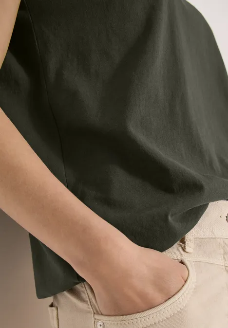 Eine Person trägt ein dunkles T-Shirt und helle Beinkleidung, Hand locker in der Hosentasche.